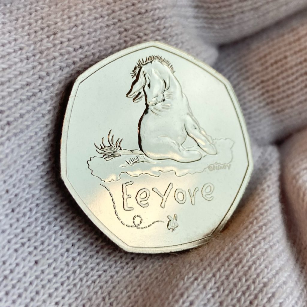 2022 UK Eeyore 50p from The Royal Mint's Winnie the Pooh 50p Collection.
Featuring a design of the original illustrations by E. H. Shephard from A. A. Milne's books.
Eeyore seen sitting on the floor, looking back at his tale on the ground.
The inscription 'Eeyore' appears, with a bumble bee beneath - the motif of the collection.