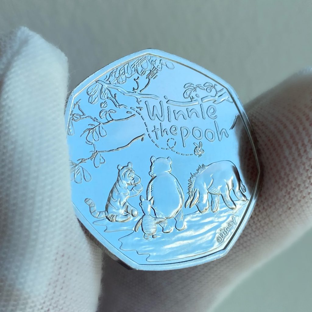 2022 UK Winnie the Pooh and Friends 50p.
Featuring design of Winnie the Pooh, Eeyore, Tigger, and Piglet all sat beneath a tree, with a bumble bee buzzing above. 
The inscription reads: 'Winnie the Pooh' and 'Disney' trademark.