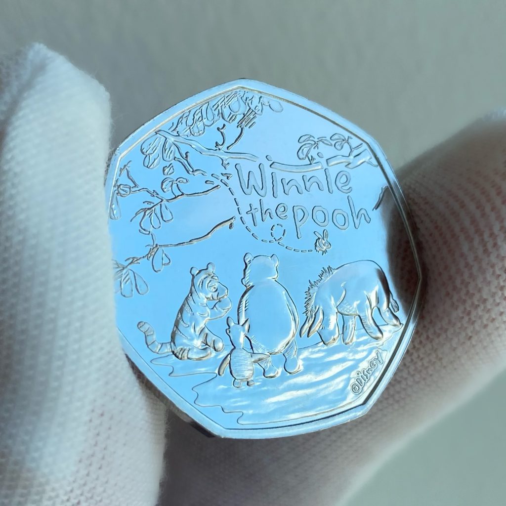 2022 UK Winnie the Pooh and Friends 50p.
Featuring design of Winnie the Pooh, Eeyore, Tigger, and Piglet all sat beneath a tree, with a bumble bee buzzing above.
The inscription reads: 'Winnie the Pooh' and 'Disney' trademark.